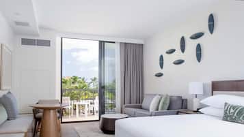 Habitación de hotel con cama, sofá, mesa de comedor y balcón con vista a palmeras.