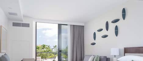 A hotel room with a bed, a sofa, a dining table, and a balcony with a view of palm trees.