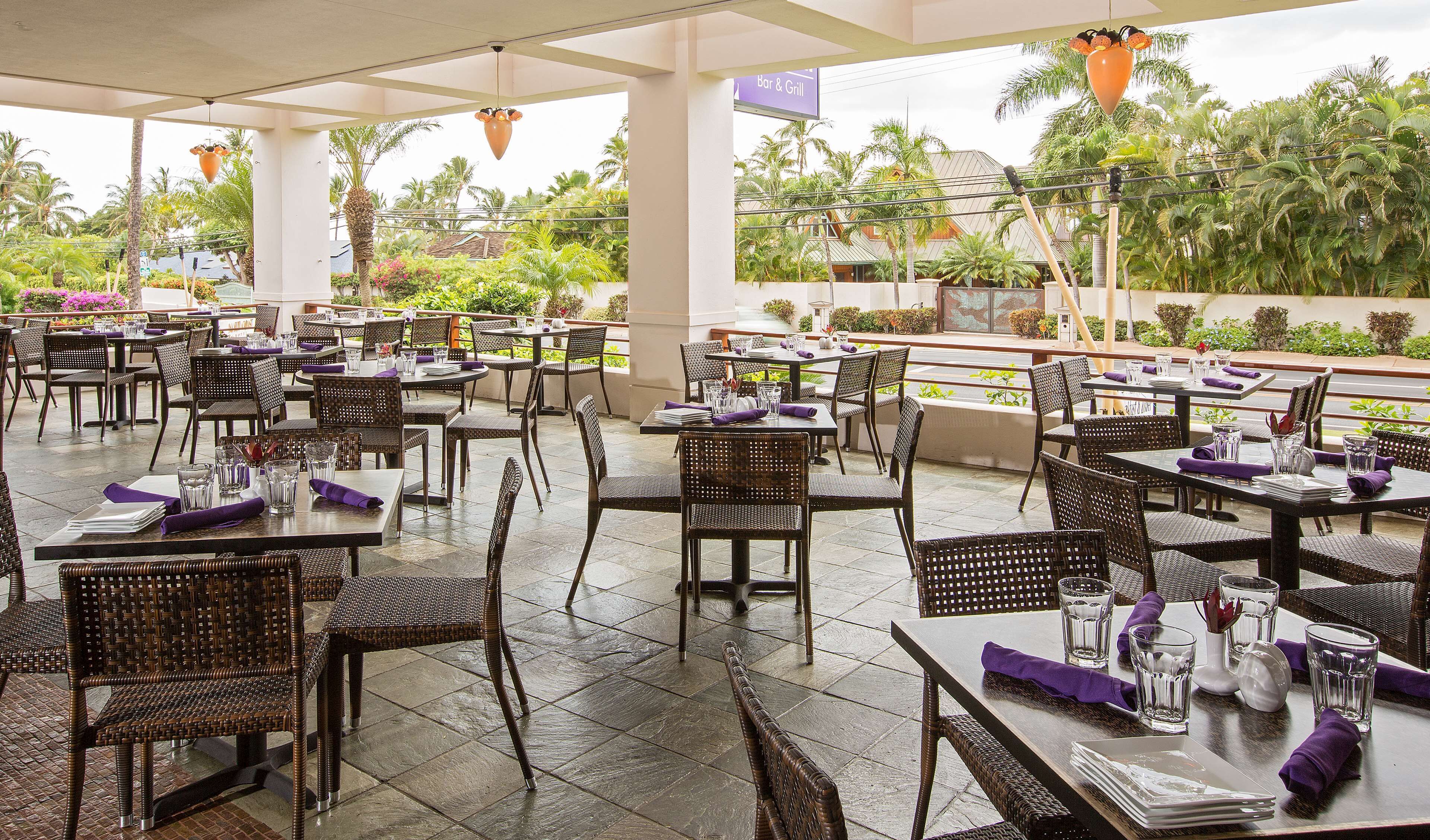 An outdoor restaurant with multiple tables and chairs, surrounded by lush greenery and palm trees.