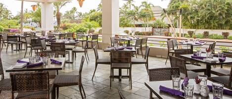 An outdoor restaurant with multiple tables and chairs, surrounded by lush greenery and palm trees.