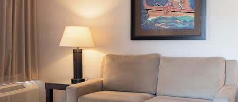 A hotel room with a beige sofa, a table lamp, a framed artwork, and a window with curtains.