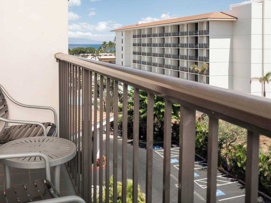 A balcony with a metal railing, a metal chair, and a small table. In the background, there is a building with multiple floors and a parking area with blue parking space markings.