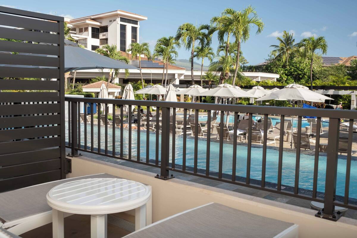 Un balcón con vista a una zona de piscina, que incluye sillones de descanso y sombrillas, y un edificio al fondo.