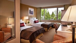 A hotel room with two beds, a desk, a chair, a lamp, a picture on the wall, and a window with a view of trees.