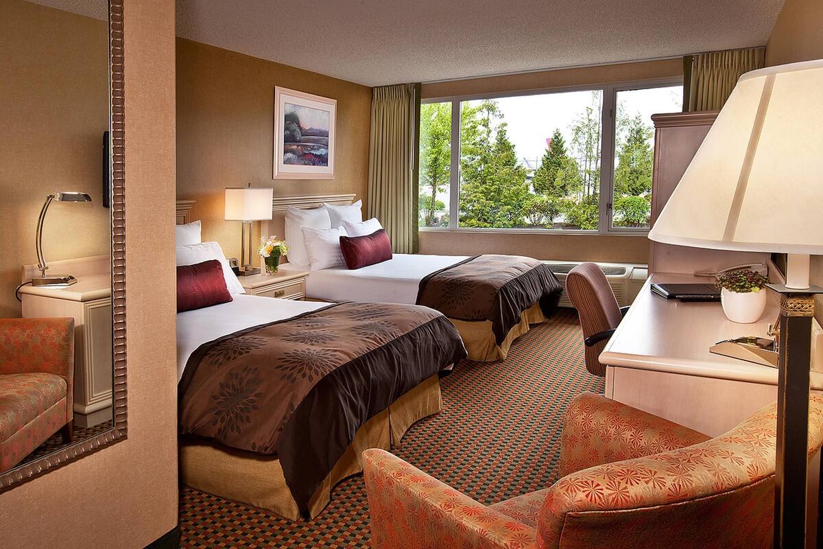 A hotel room with two beds, a desk, a chair, a lamp, a picture on the wall, and a window with a view of trees.