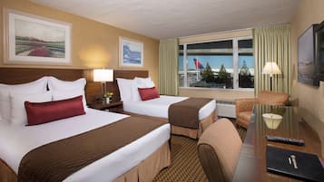A hotel room with two beds, a desk, a chair, a window with a view of an airport, and a lamp.