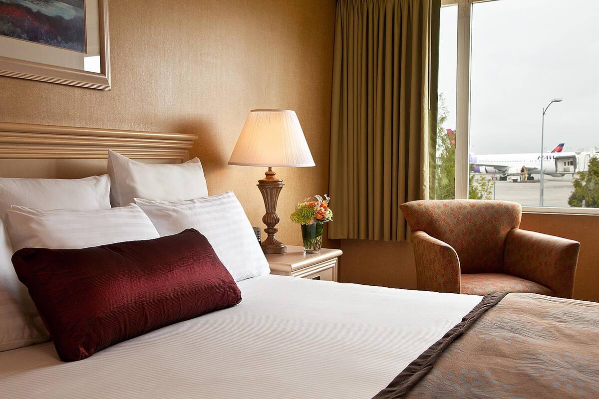 A hotel room with a large bed, a nightstand with a lamp, a chair, and a window with a view of an airport.