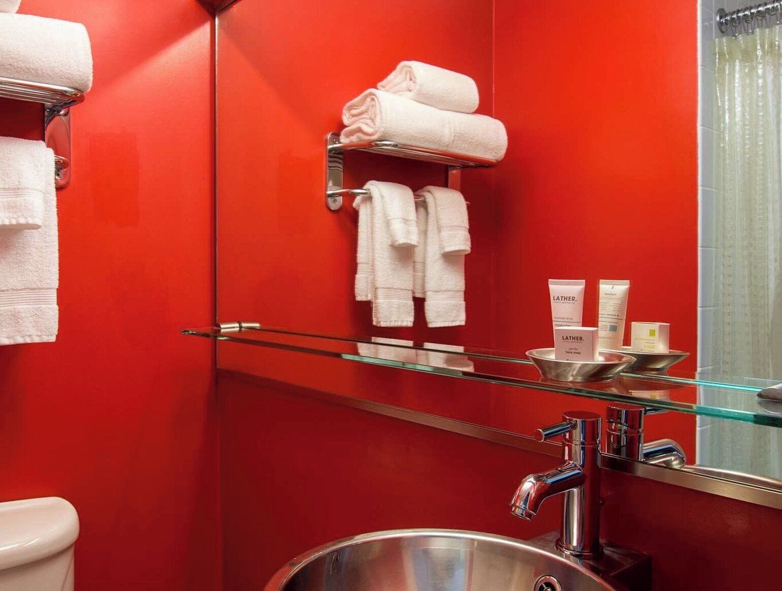 A bathroom with a red wall, a glass shelf with toiletries, and a mirror.