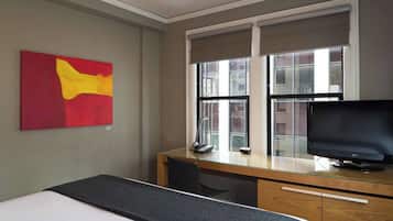 A hotel room with a bed, a desk with a computer and phone, a TV, and a window with blinds.