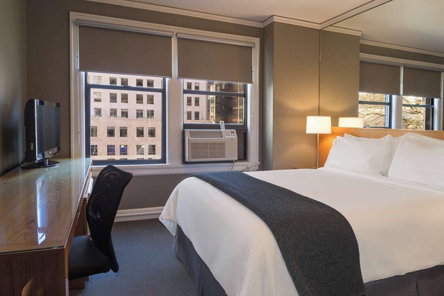 A hotel room with a bed, a desk with a computer, a chair, a window with blinds, and a lamp.