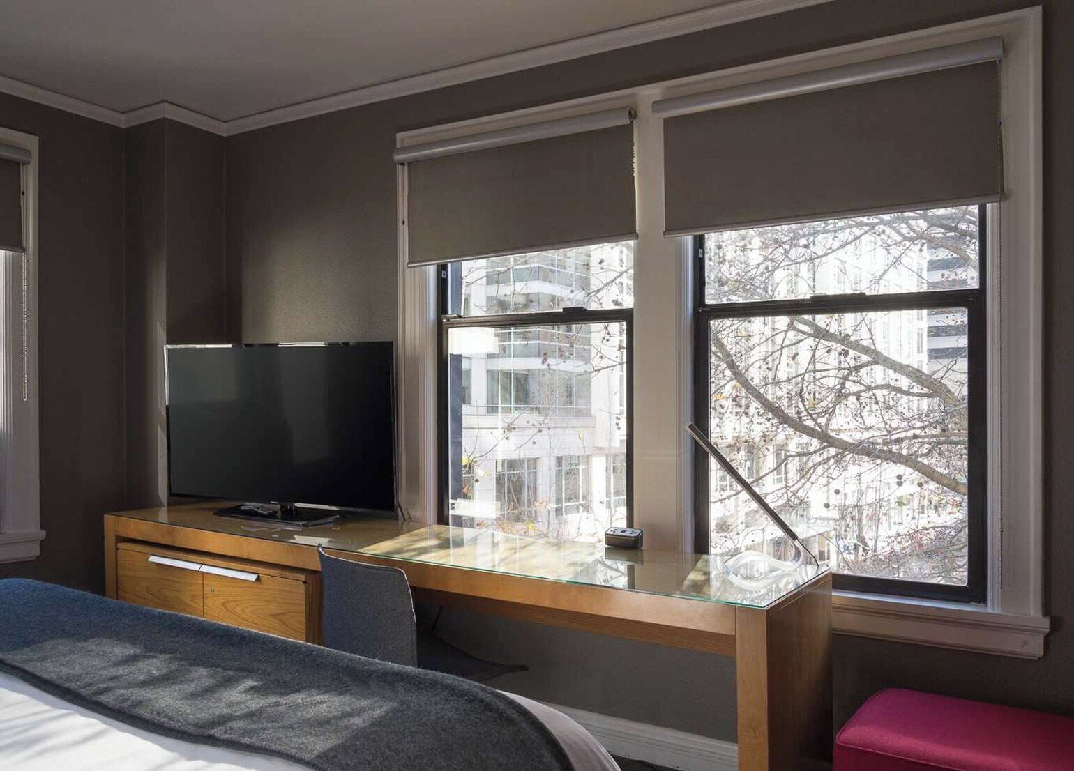 A hotel room with a bed, a desk with a TV, and large windows with a view of buildings and trees.