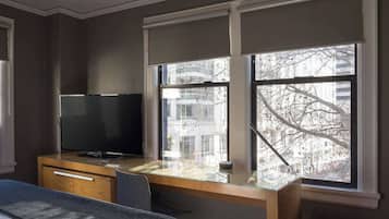 A hotel room with a bed, a desk with a TV, and large windows with a view of buildings and trees.