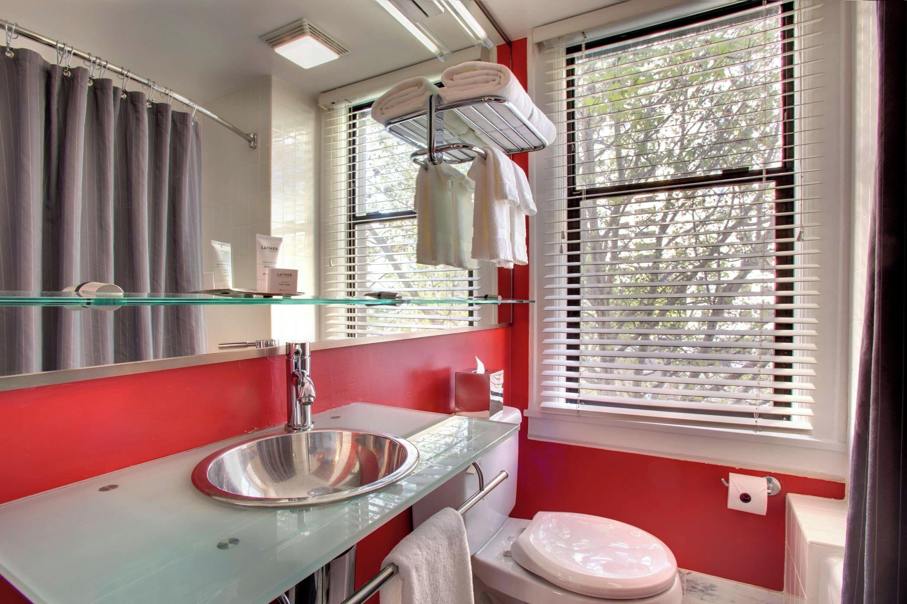 A bathroom with a glass sink, a mirror, a toilet, and a window with blinds.