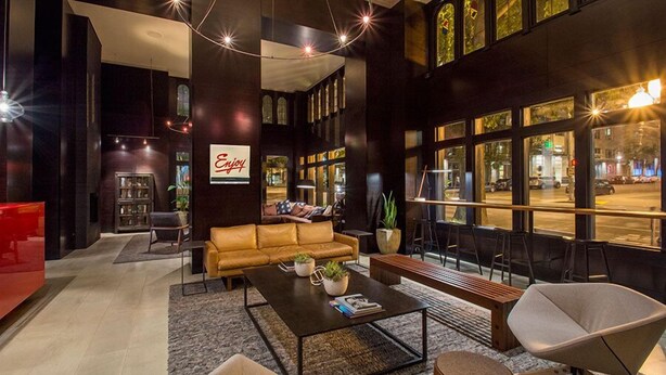 A modern lobby with a leather sofa, a coffee table, and a red accent chair. There is a bookshelf and a sign that says "Enjoy". - Hotel Max (Seattle)