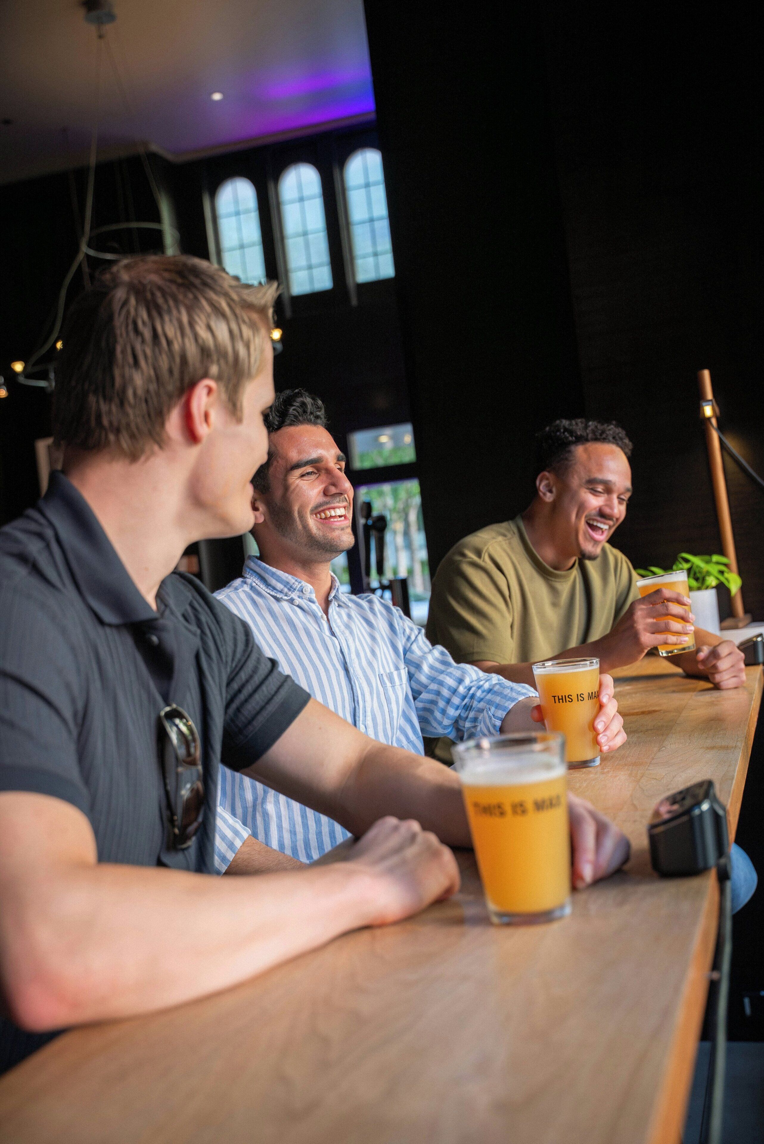 A group of people seated at a bar with drinks, in a room with large windows and a modern interior.