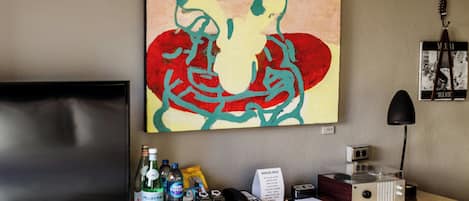 A hotel room with a flat-screen TV, a desk with a telephone, a coffee machine, and a wall-mounted artwork.