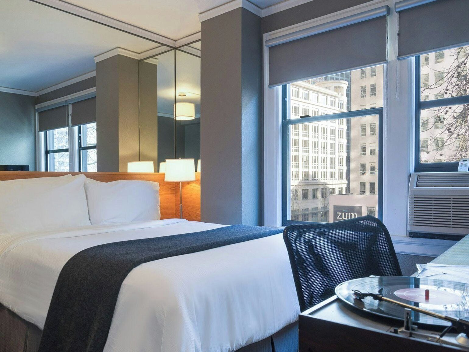 Habitación de hotel con cama, lámparas de noche, una silla, una mesa con bandeja y ventana con vista a la ciudad.