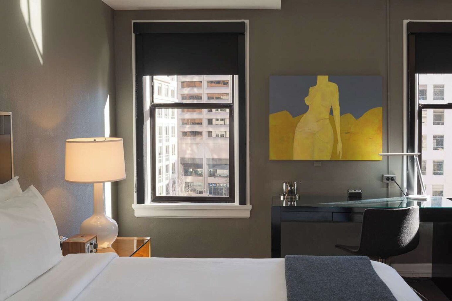 A hotel room with a bed, a desk, a lamp, a painting, and a window with a city view.