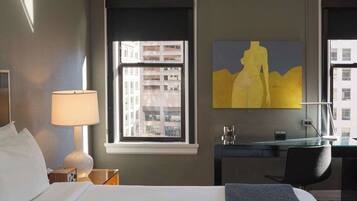 A hotel room with a bed, a desk, a lamp, a painting, and a window with a city view.