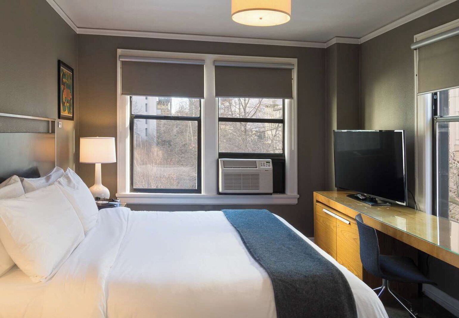 A hotel room with a bed, a desk with a television, a window with blinds, and an air conditioning unit.