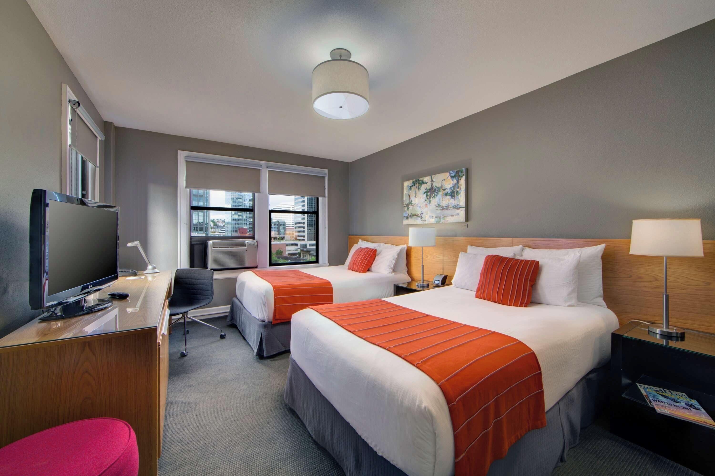 A hotel room with two beds, a desk with a computer, a TV, and a window with blinds.