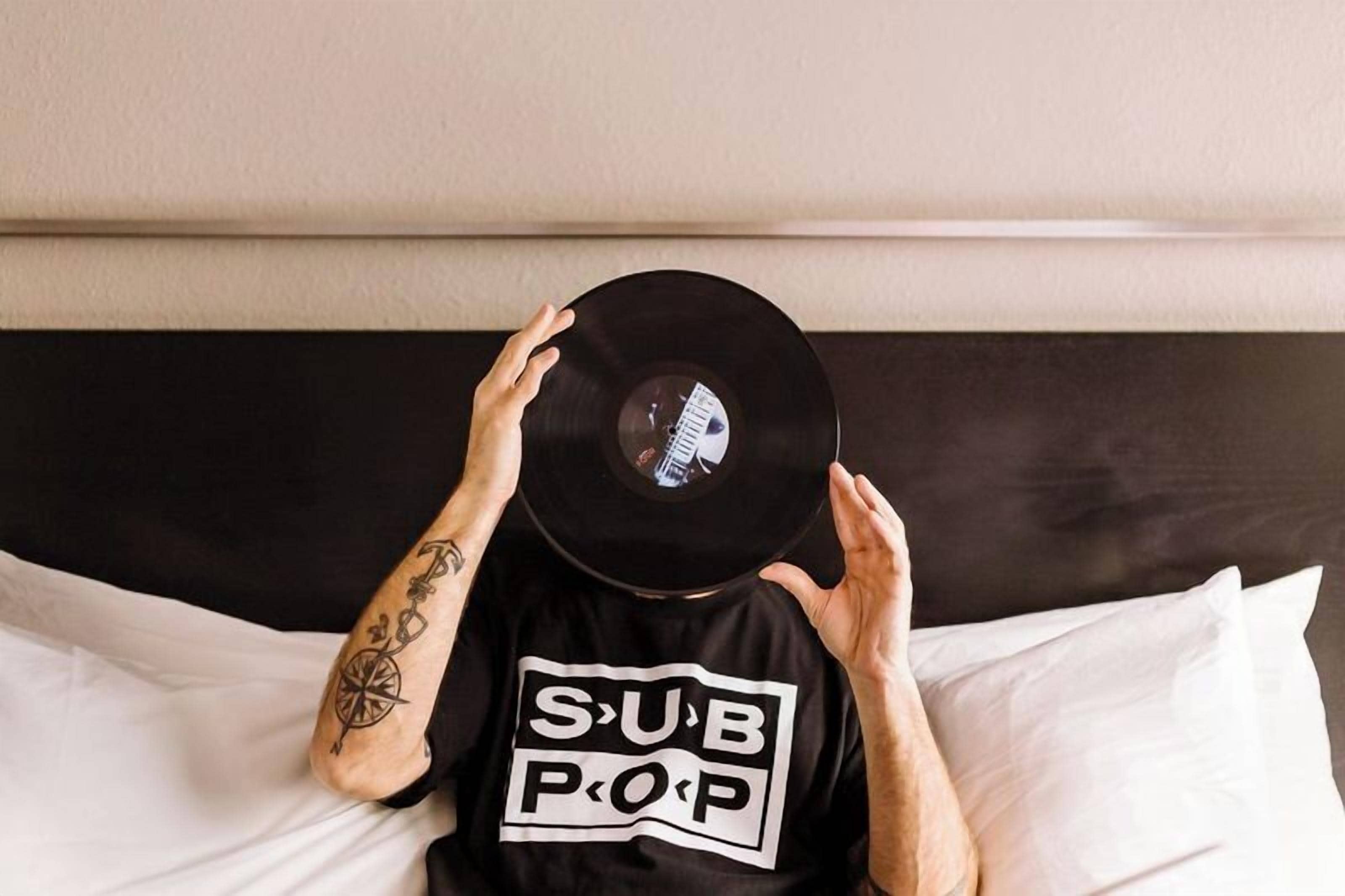 Une personne allongée sur un lit dont la tête de lit est noire, portant un t-shirt noir avec un texte blanc.
