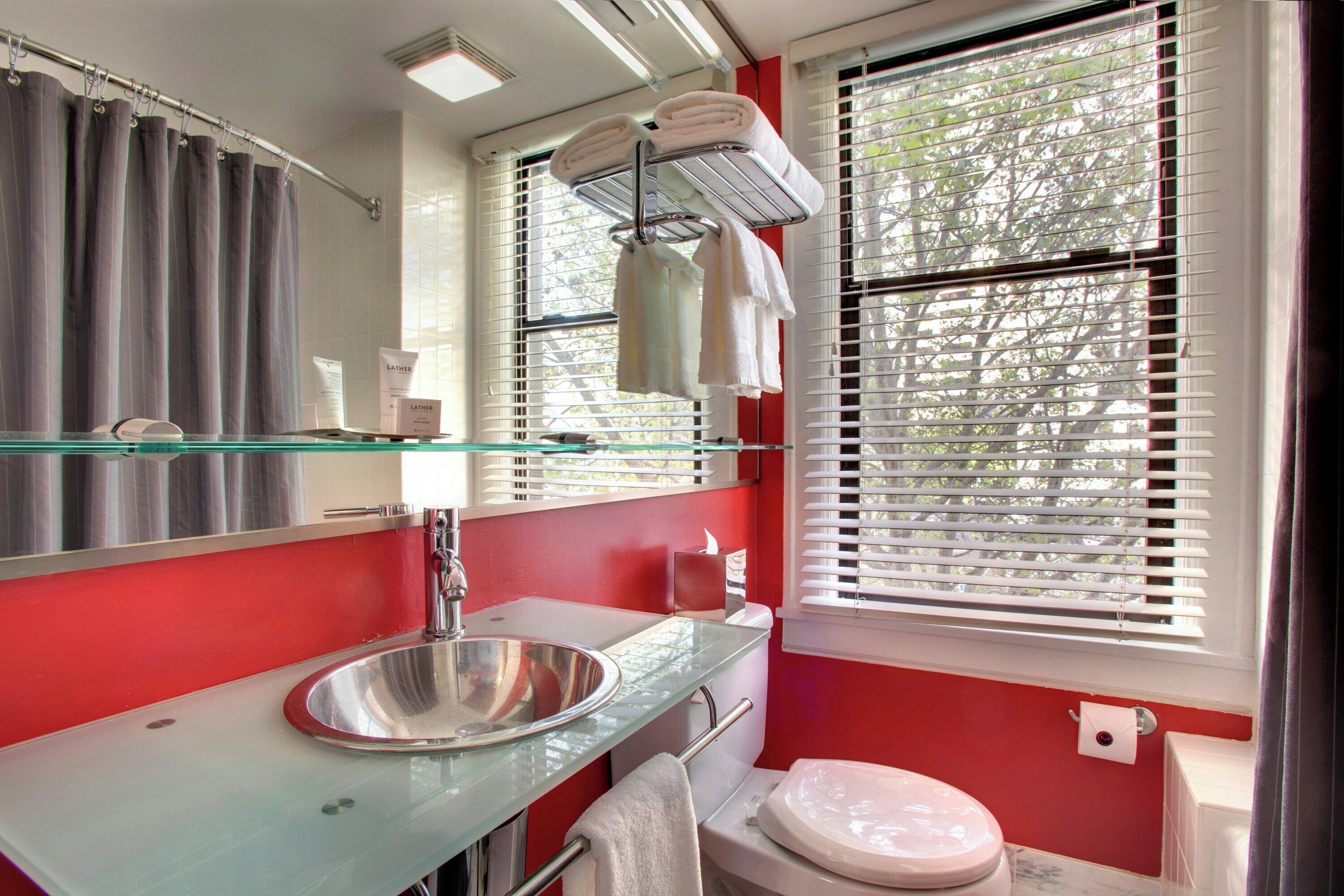 A bathroom with a glass sink, a mirror, a toilet, and a window with blinds.