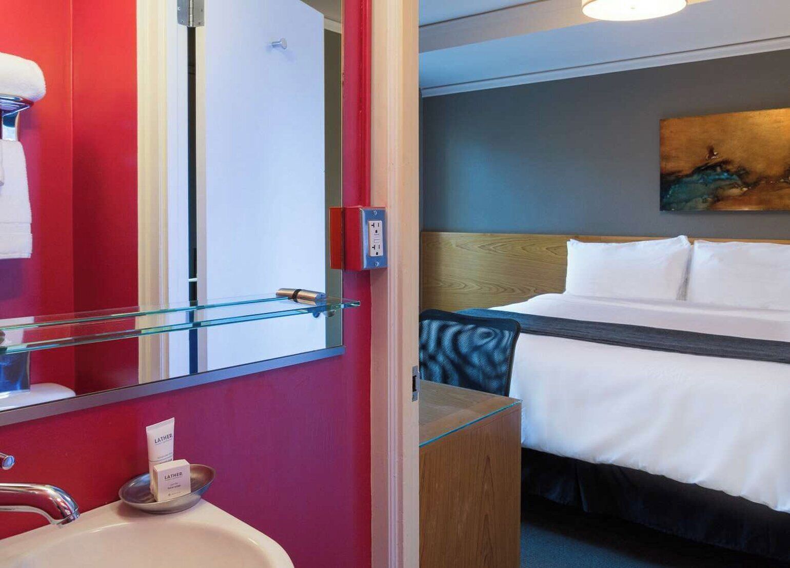 A hotel room with a bed, a bedside table, a mirror, a sink, and a towel rack.