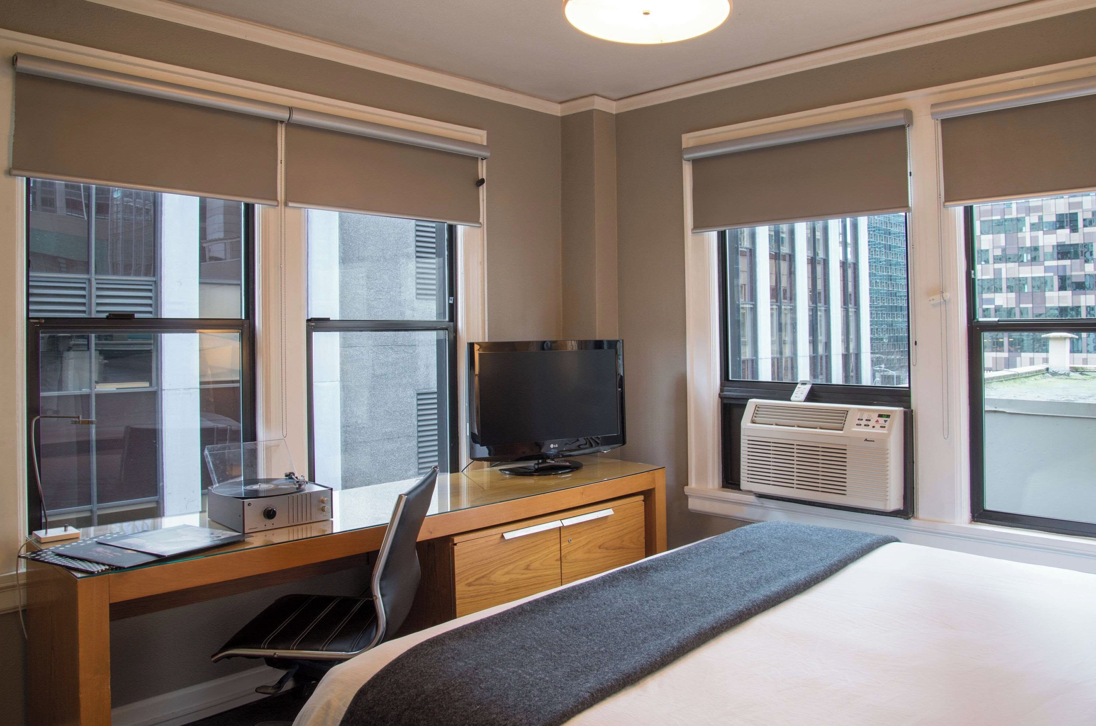 A hotel room with a bed, a desk with a computer and a flat-screen TV, a chair, and an air conditioning unit.