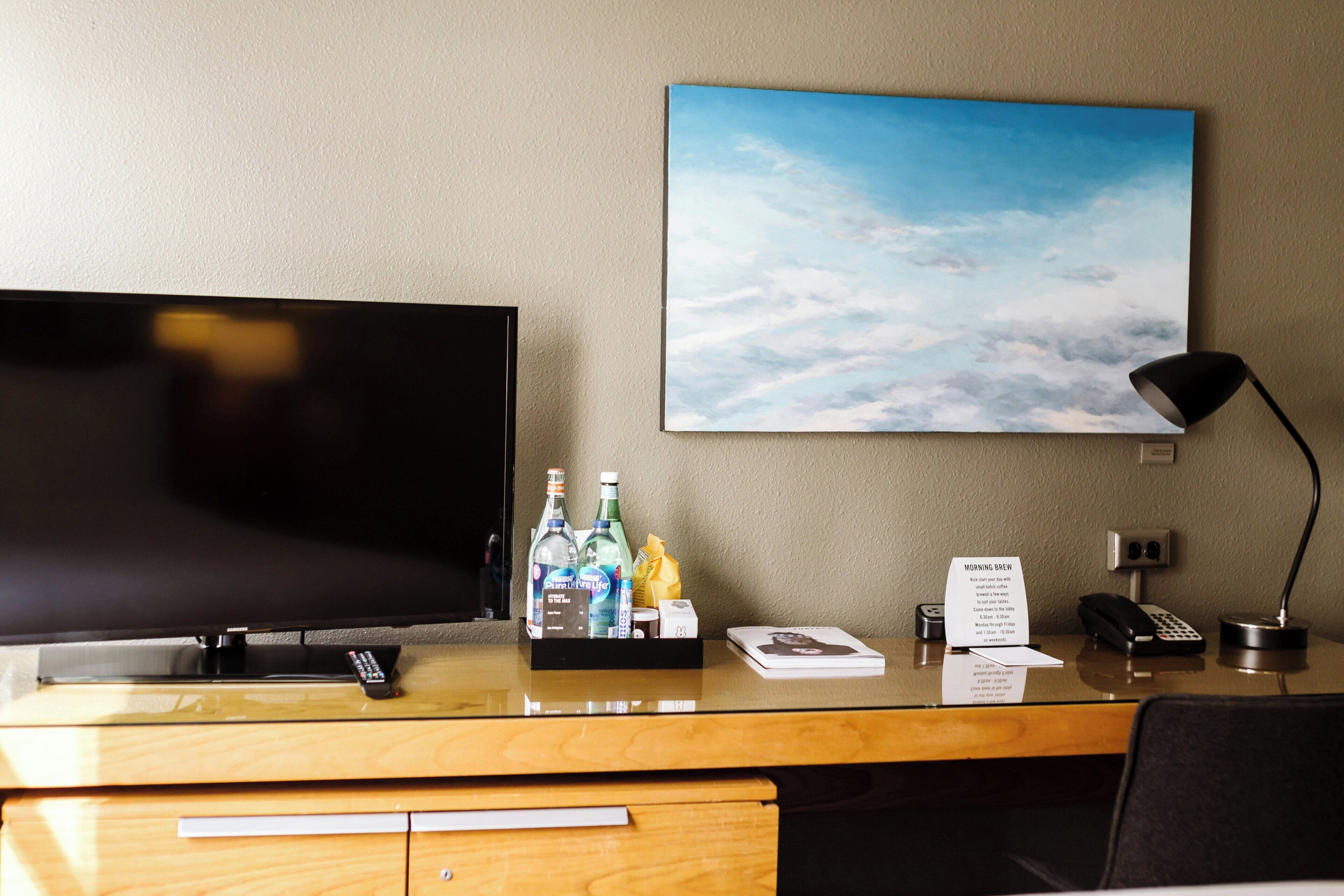 Habitación de hotel con televisión, escritorio con lámpara, una botella de agua, control remoto y un cuadro de nubes.