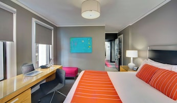 A hotel room with a bed, desk, chair, and a painting on the wall.
