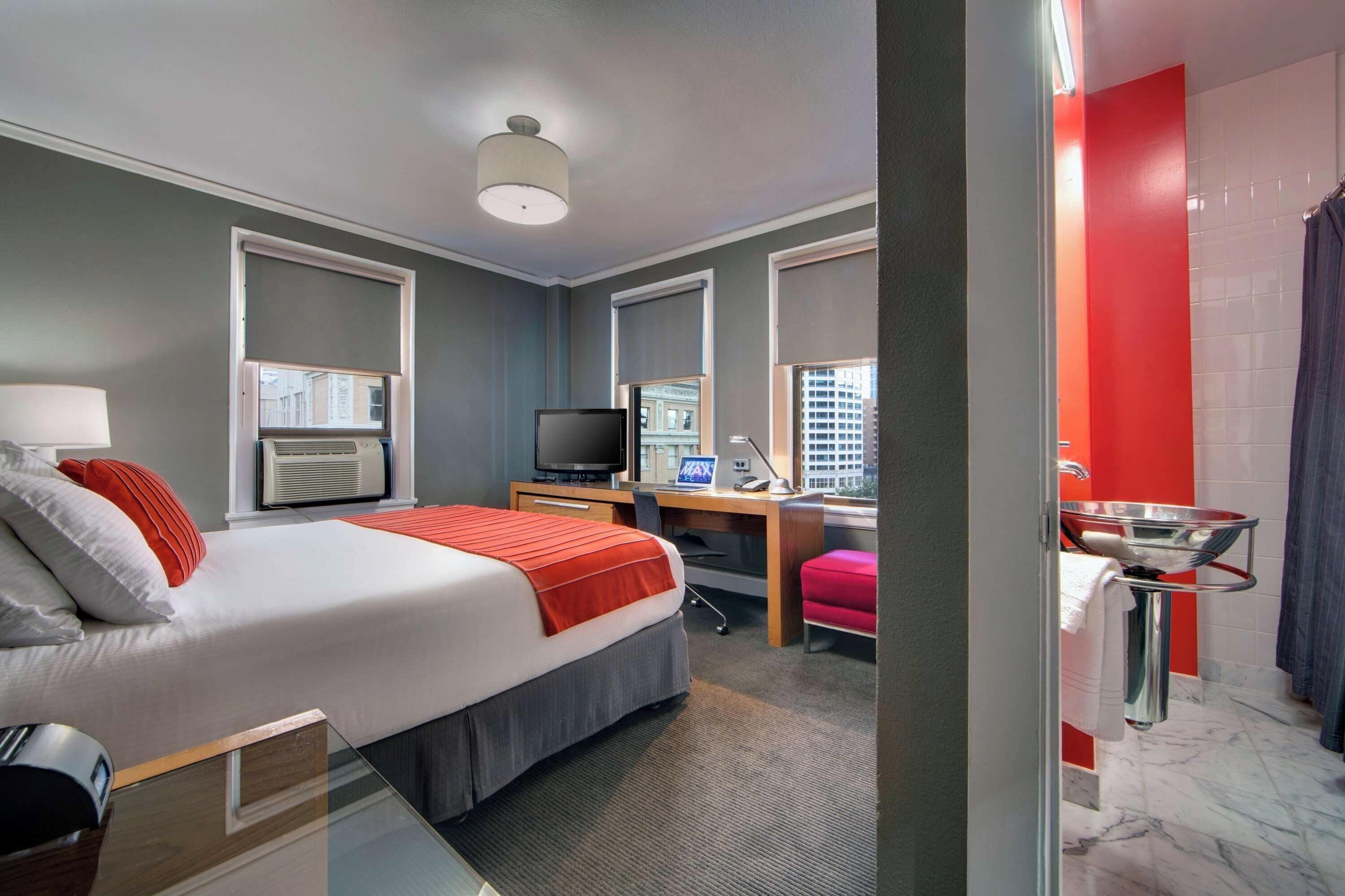 Habitación de hotel con cama, escritorio, televisión y baño con ducha y lavamanos.