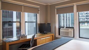 A hotel room with a bed, a desk with a computer and a television, and large windows with blinds.