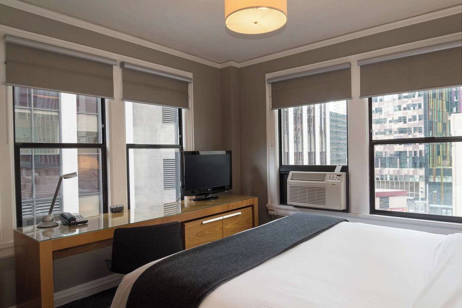 A hotel room with a bed, a desk with a television, a chair, and large windows with city views.