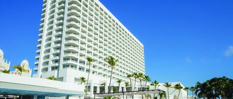 A large hotel with a swimming pool area, palm trees, and a clear blue sky.