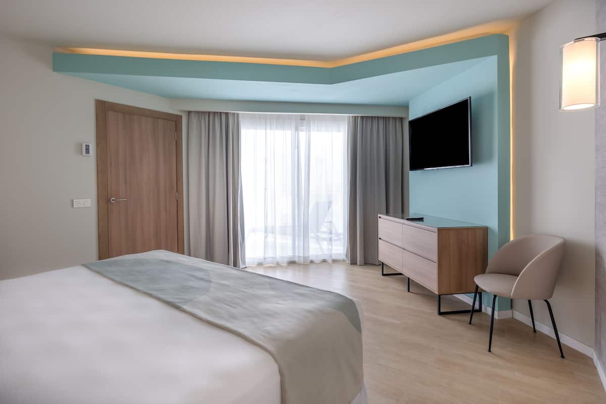 A modern hotel room with a large bed, a desk, a chair, and a TV mounted on the wall.