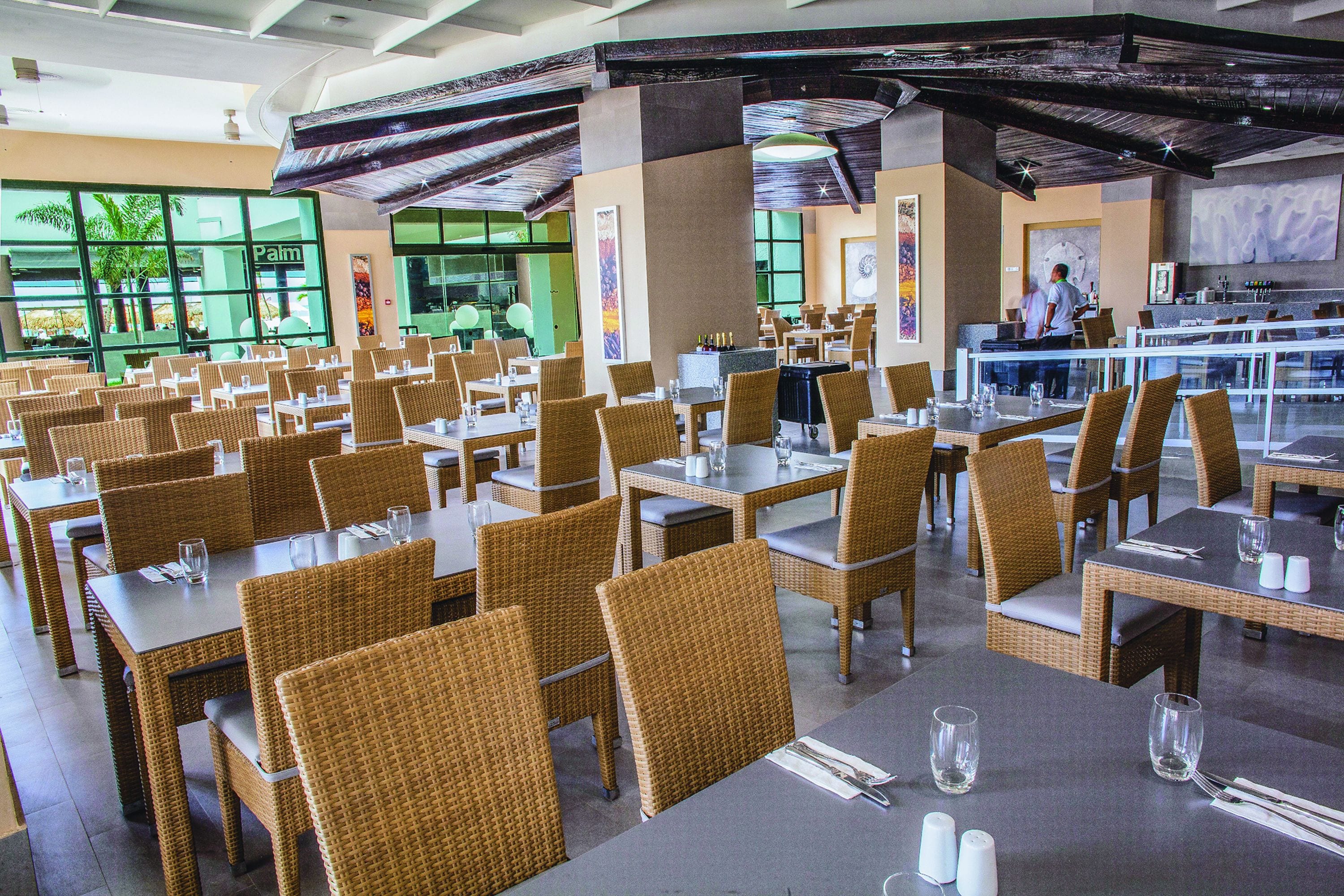A spacious restaurant with wicker chairs and tables, large windows, and a high ceiling with exposed beams.