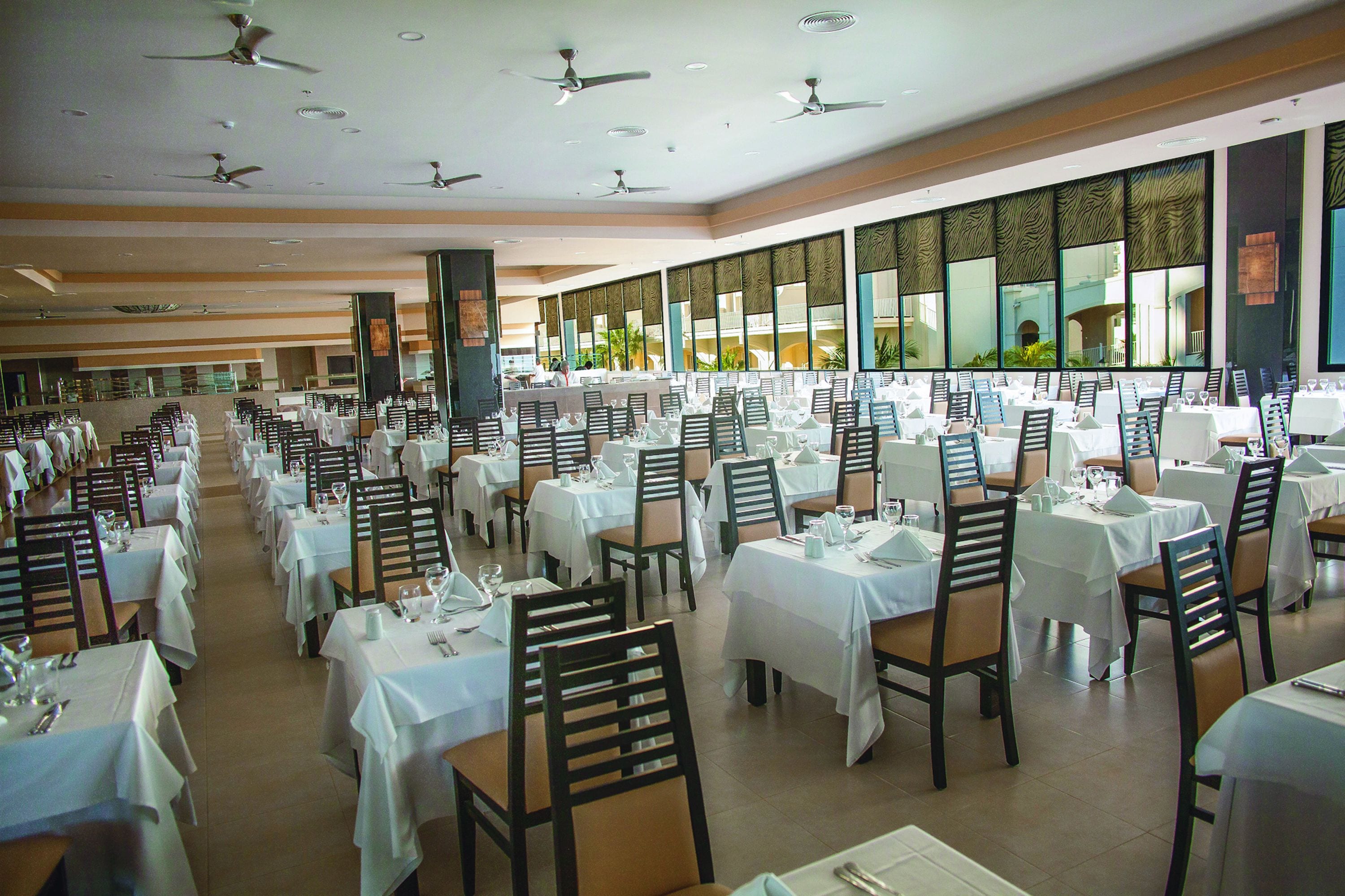 A large dining hall with multiple tables and chairs, ceiling fans, and large windows with decorative panels.