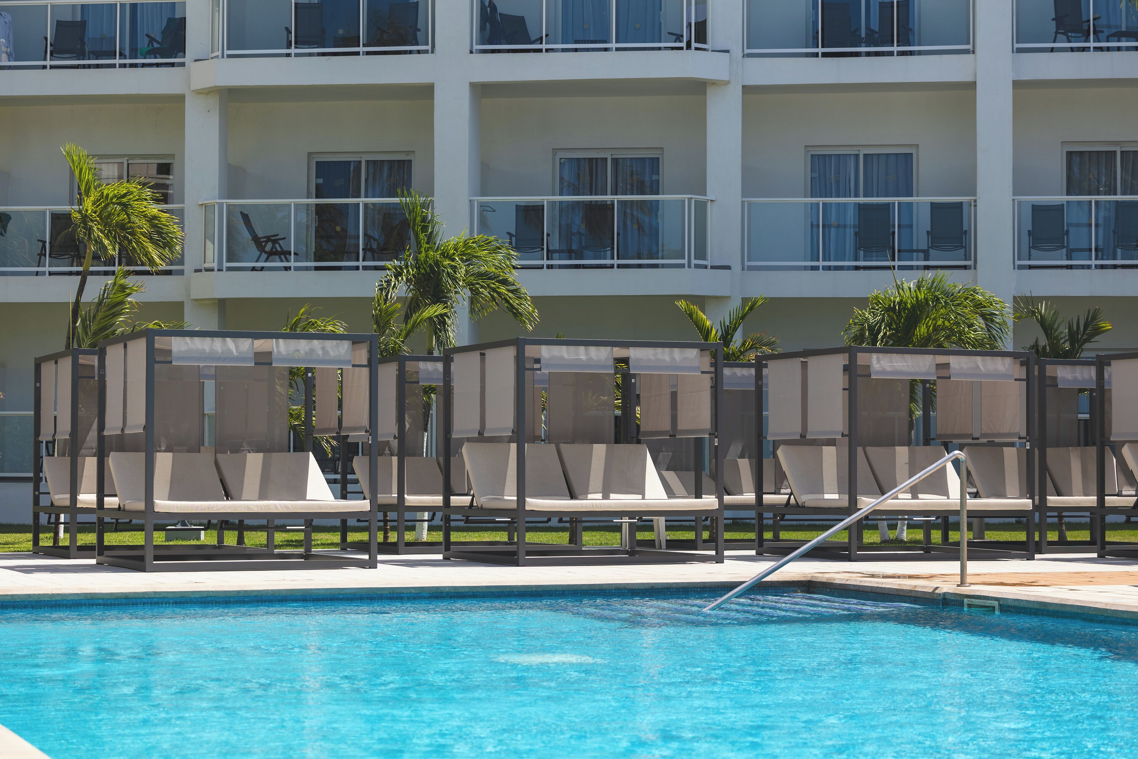 Zona de piscina con sillas de descanso y palmeras, rodeada por un edificio de varios pisos con balcones.