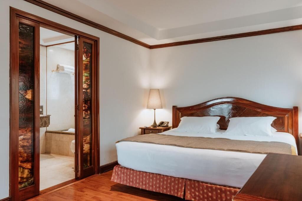 Una habitación de hotel con una cama de madera, una mesita de noche con lámpara, un espejo y piso de madera.