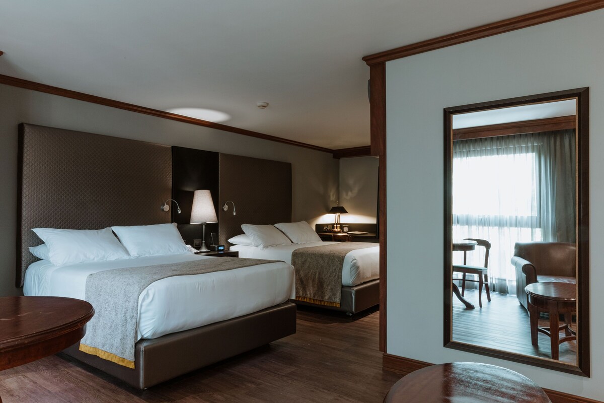 A hotel room with two beds, a large mirror, a wooden floor, and a view of the outside through the window.