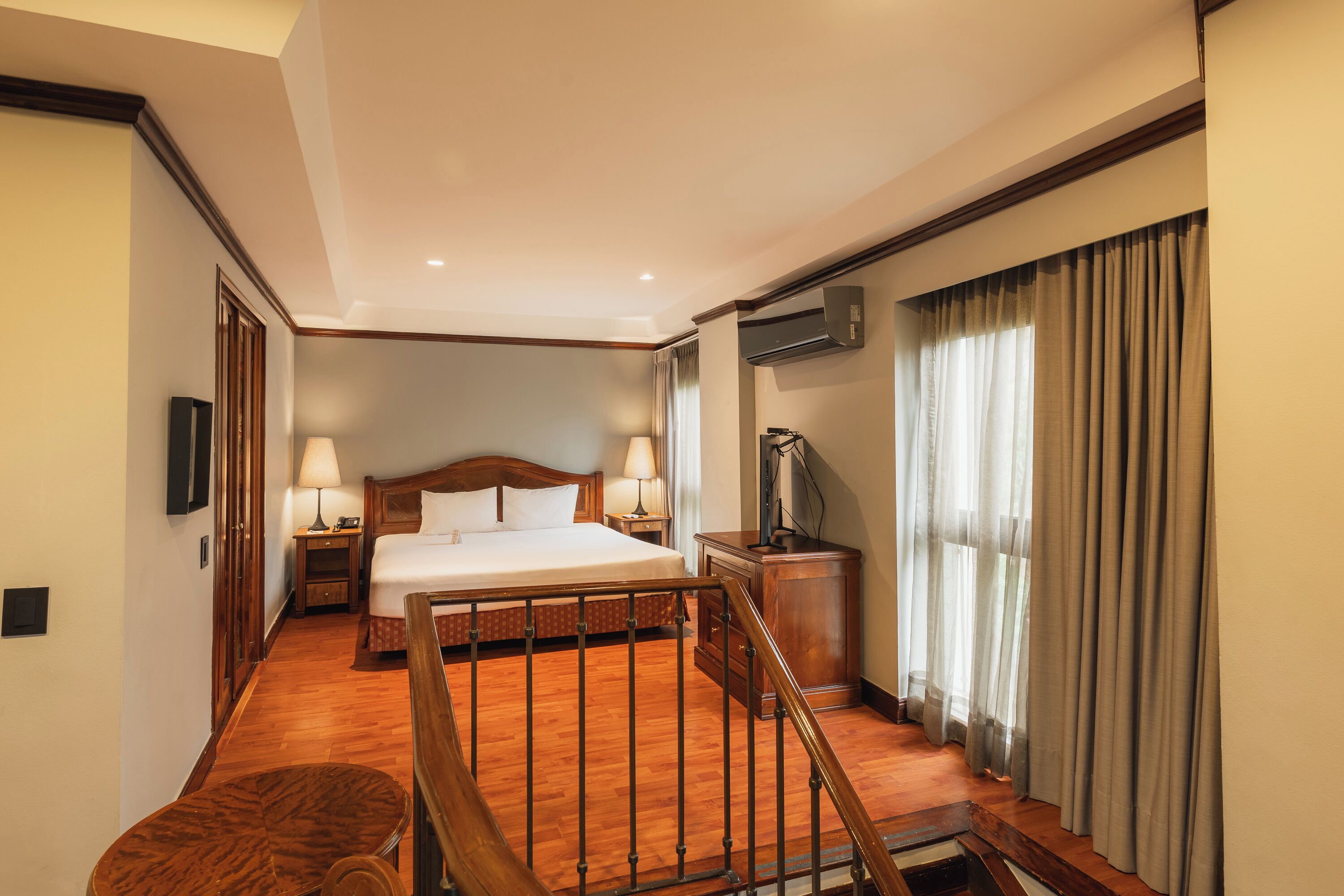 A hotel room with a wooden bed, a desk, a chair, a TV, and a window with curtains.