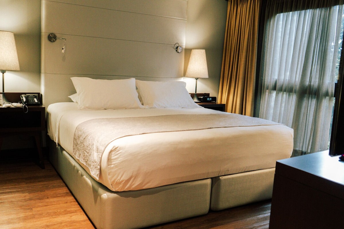 A hotel room with a large bed, two bedside tables with lamps, a nightstand, a dresser, and a window with curtains.