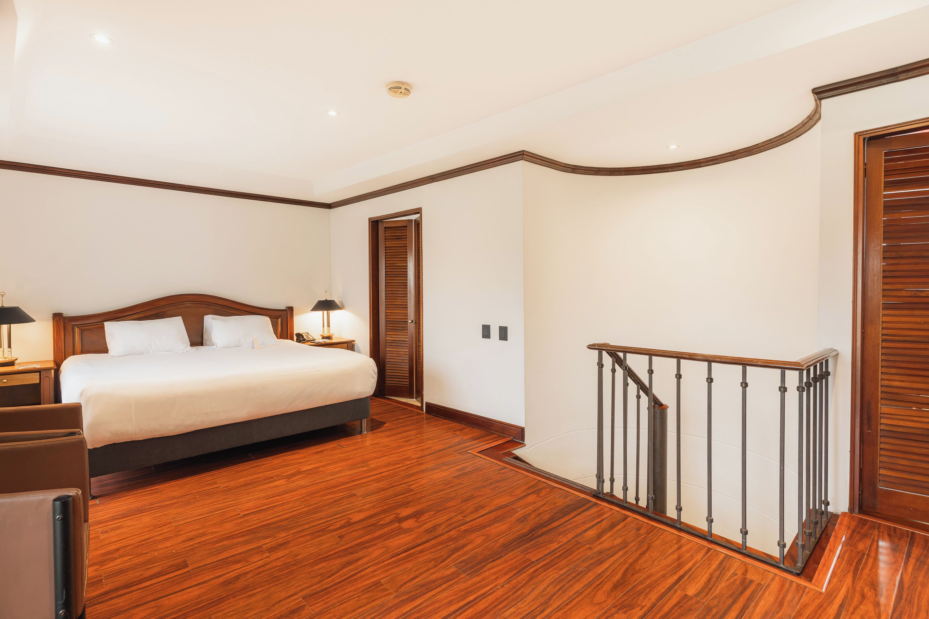 A hotel room with a wooden floor, a bed with white linens, a nightstand with a lamp, and a staircase with a metal railing.
