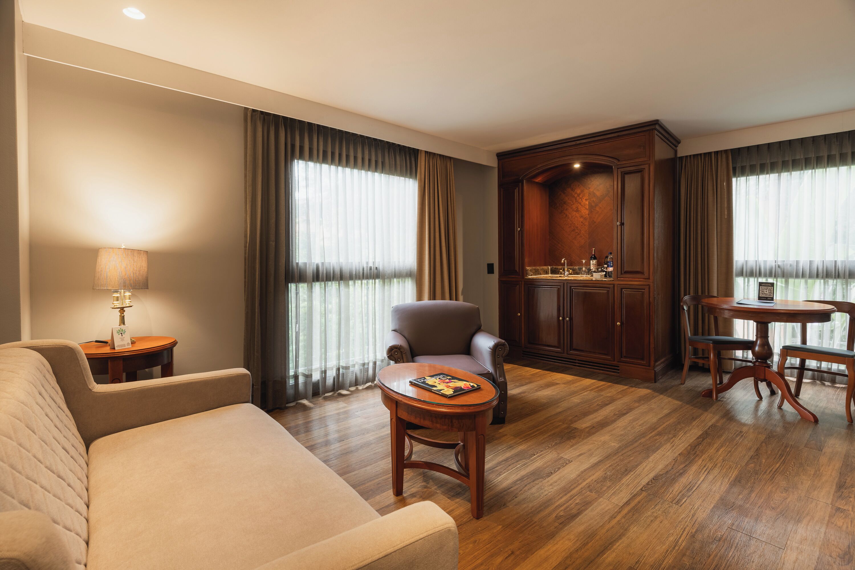 A hotel room with a sofa, a small round table with a magazine, a cabinet, and a dining table with chairs.