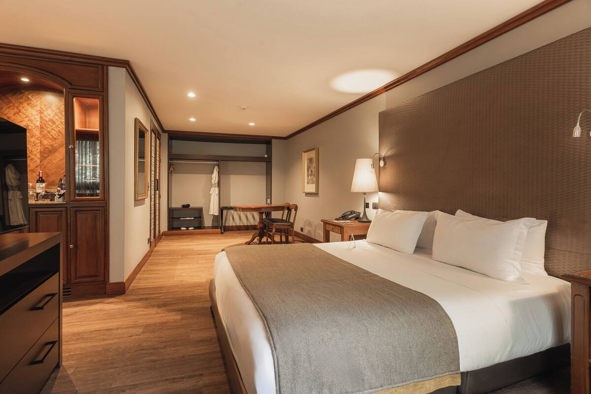 A hotel room with a large bed, a desk, a chair, a lamp, and a wardrobe.