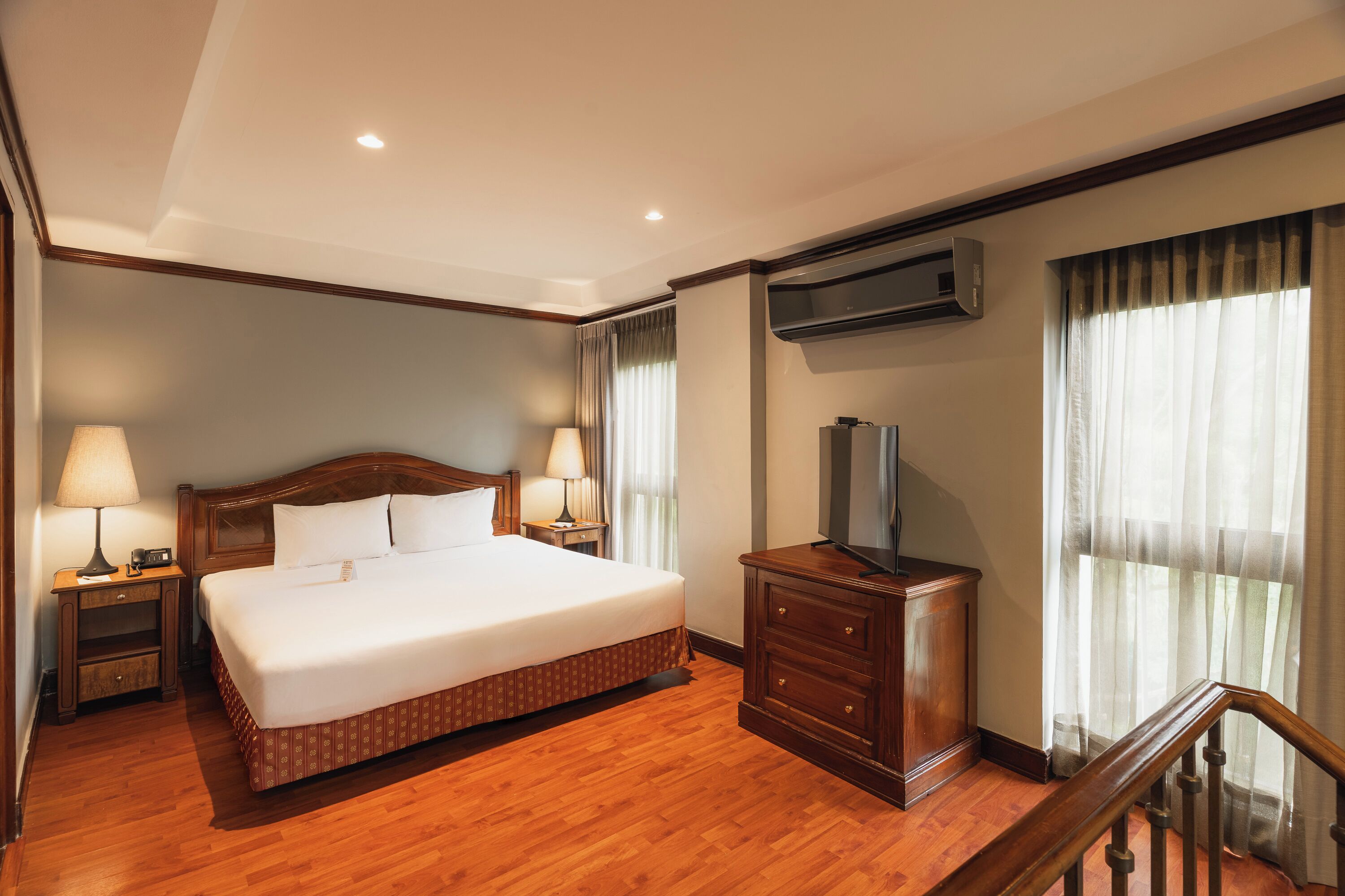 A hotel room with a large bed, wooden flooring, a dresser, a TV, and an air conditioning unit.
