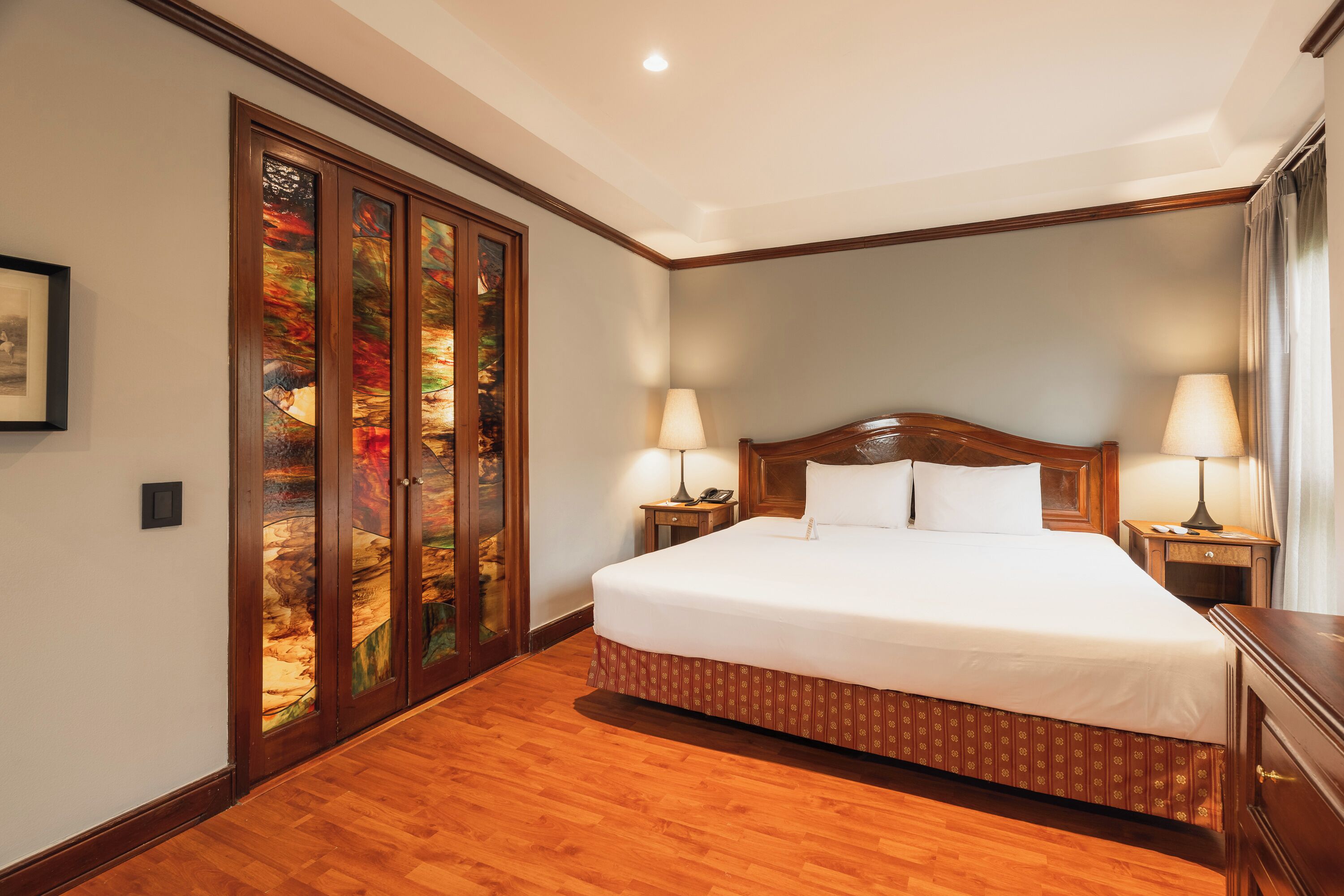 A hotel room with a large bed, wooden floor, two bedside tables with lamps, and a large window with curtains.