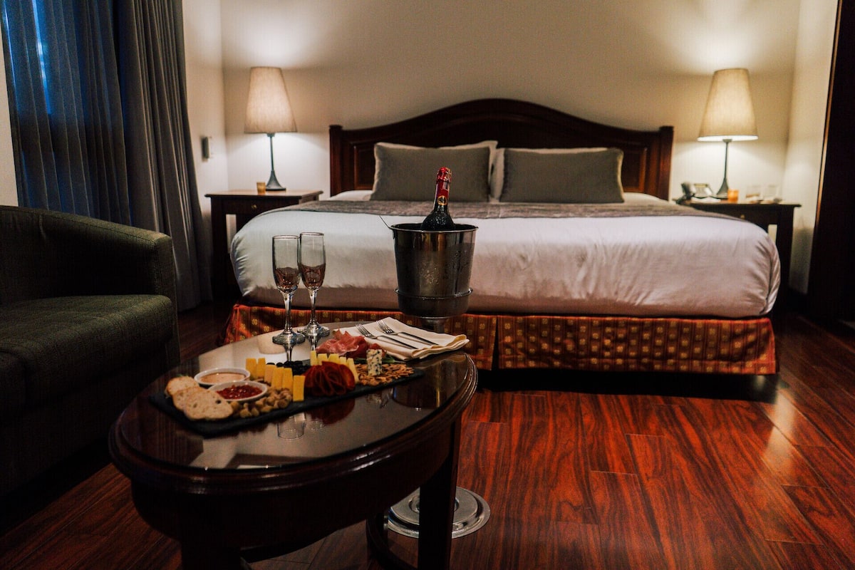 A hotel room with a bed, bedside tables, a dining table with food, and a chair.