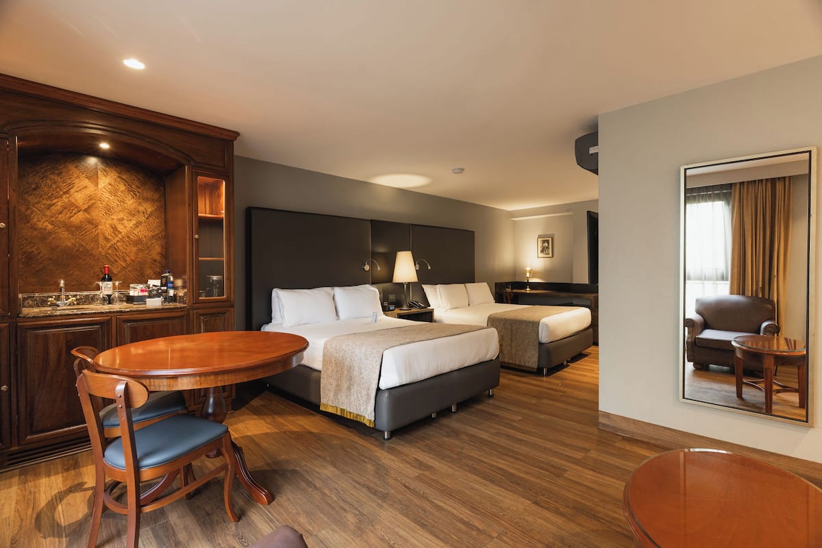 A hotel room with a wooden dining table, two beds, a sofa, a small table, and a mirror.